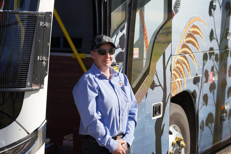 A female bus driver stands by the open door of a coach covered in an Aboriginal artwork.